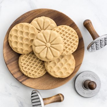 Golden baked cookies with decorative stamped patterns arranged on white plate beside three Nordic Ware cookie stamps