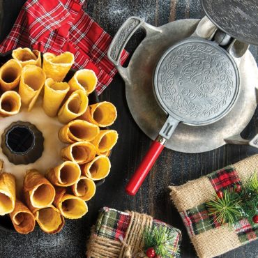 Golden krumkake cookie cones arranged in Nordic Ware bundt pan with open aluminum krumkake iron showing detailed plate pattern