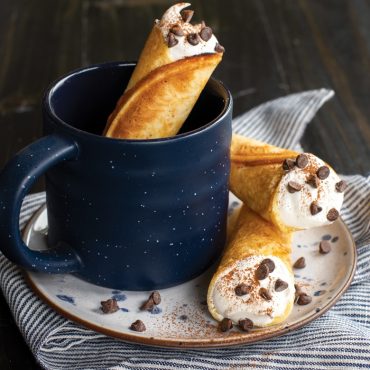 Three golden krumkake cookies filled with whipped cream and chocolate chips, two on white plate, one shaped as cup