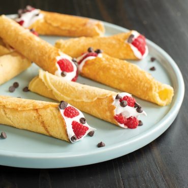 Golden cone-shaped krumkake filled with whipped cream, fresh raspberries and chocolate chips on white plate