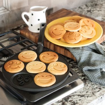 Nordic Ware aluminum smiley face pancake pan on stovetop with six golden cooked pancakes showing happy face designs