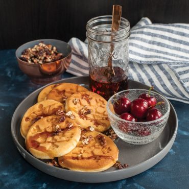 Six golden snowflake-shaped pancakes on white plate with syrup, chopped nuts, and fresh cherries
