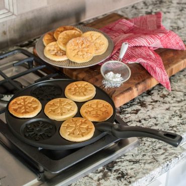 Nordic Ware aluminum pancake pan on stovetop with seven golden snowflake-shaped pancakes, detailed fluted design
