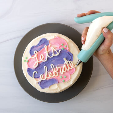 Hand using Nordic Ware E-Z Deco icing pen to pipe white frosting decorations on round white frosted cake