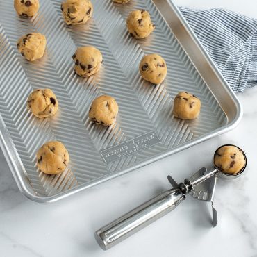 Silver stainless steel cookie scoop holding chocolate chip cookie dough with unbaked dough balls on baking sheet