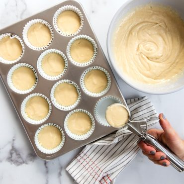 Nordic Ware large cookie scoop filling muffin tin with vanilla batter, paper liners in aluminum cavities