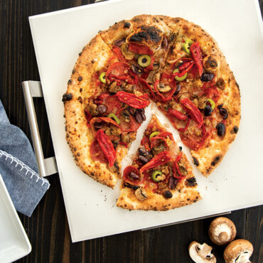 Baked pizza with golden crust and melted cheese on Nordic Ware square stone pizza stone, one slice lifted