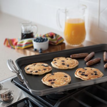 Nordic Ware aluminum two-burner griddle with raised sides on stovetop, cooking golden pancakes and browned sausage links
