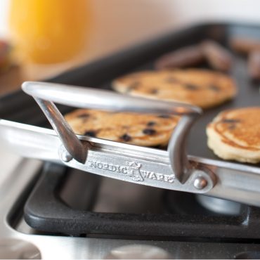 Nordic Ware aluminum two-burner griddle with raised sides on stovetop, golden pancakes cooking on surface