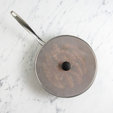 Nordic Ware 13-inch mesh spatter cover with stainless steel handle covering skillet of cooking bacon