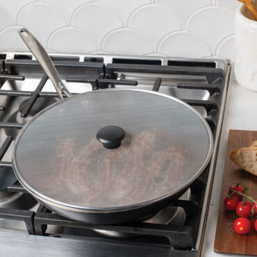 Nordic Ware 13-inch aluminum spatter cover with black knob on gas stove, covering skillet with sizzling bacon