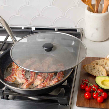 Nordic Ware 13-inch glass spatter cover partially covering skillet with sizzling bacon strips