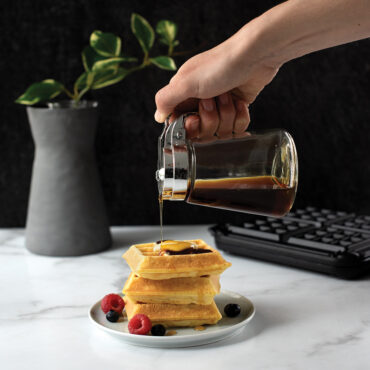 Golden Belgian waffles stacked on white plate with maple syrup being poured, Nordic Ware waffler visible