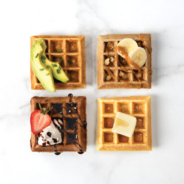 Four golden Belgian waffle squares with varied toppings: avocado, banana with nuts, chocolate with strawberry, and butter pats