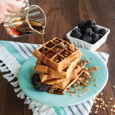 Golden Belgian waffles stacked on mint green plate with maple syrup being poured from glass pitcher, fresh blackberries
