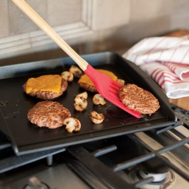 Nordic Ware non-stick backsplash griddle with seared burgers, mushrooms, and red silicone spatula on stovetop