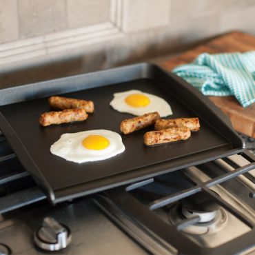 Nordic Ware non-stick backsplash griddle with fried eggs and sausage links cooking on stovetop