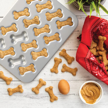 Golden-brown bone-shaped dog treats baked in Nordic Ware aluminum puppy-themed baking pan with additional treats scattered nearby