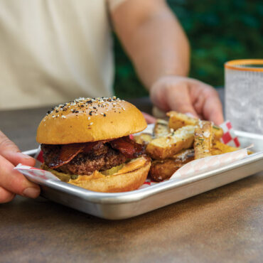 White rectangular serving tray holding sesame seed burger, loaded fries, and pickles being held outdoors