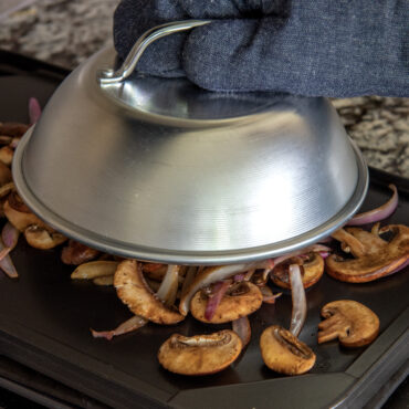 Nordic Ware aluminum cheese melting dome covering colorful grilled vegetables on griddle surface