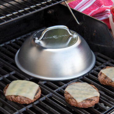Nordic Ware aluminum cheese melting dome covering hamburgers on grill, silver finish with black handle