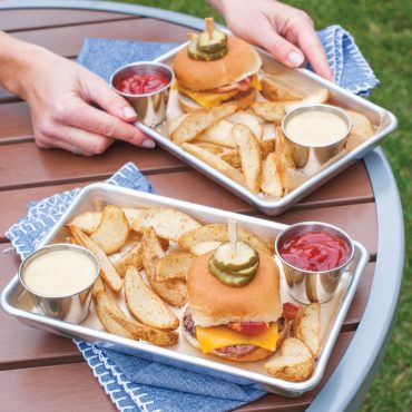 Two Nordic Ware aluminum burger serving trays holding cheeseburgers, potato chips, and dipping sauces outdoors