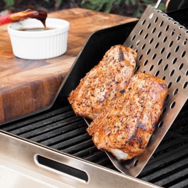 Two seasoned pork chops cooking under Nordic Ware perforated aluminum grill tent on outdoor grill grates