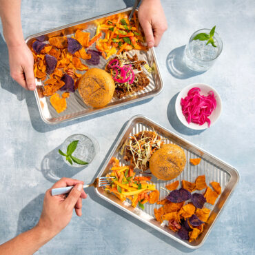 Two Nordic Ware aluminum prep trays holding grilled burgers, golden fries and chips in outdoor BBQ setting