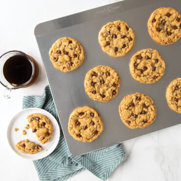 Golden baked chocolate chip cookies on Nordic Ware aluminum insulated baking sheet, one cookie cut in half on plate
