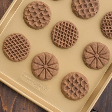 Nordic Ware nonstick baking sheet with eight baked chocolate cookies featuring decorative stamped patterns