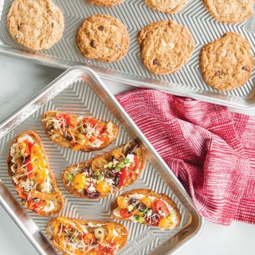 Two Nordic Ware aluminum prism baking sheets with golden baked bruschetta and cookies, textured non-stick surface
