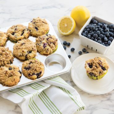 Nordic Ware aluminum muffin pan with 12 golden blueberry muffins, one served on white plate with fresh berries