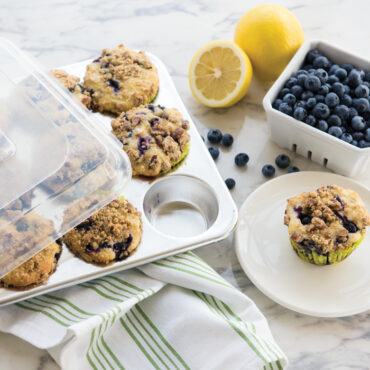 Nordic Ware aluminum 12-cup muffin pan with clear plastic storage lid, containing golden-brown blueberry muffins