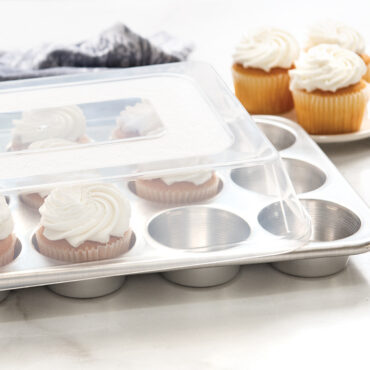 Nordic Ware aluminum 12-cavity muffin pan with clear high domed lid, containing golden vanilla cupcakes