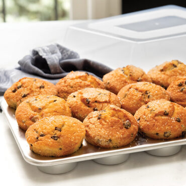 Nordic Ware aluminum muffin pan with 12 golden chocolate chip muffins, high domed lid in background