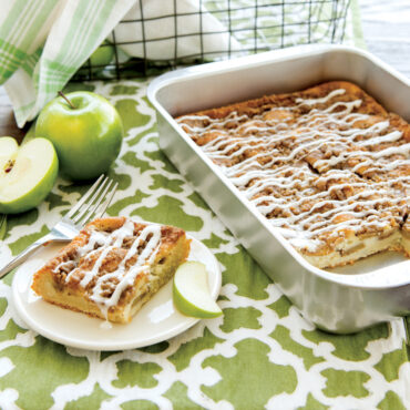 Nordic Ware aluminum 9x13 baking pan with apple streusel cake, white icing drizzle, slice plated beside fresh apples