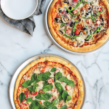 Two Nordic Ware aluminum pizza pans with baked pizzas - one traditional smooth, one perforated crisper pan