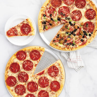 Two Nordic Ware aluminum pizza pans with baked pizzas - one traditional smooth, one perforated crisper pan