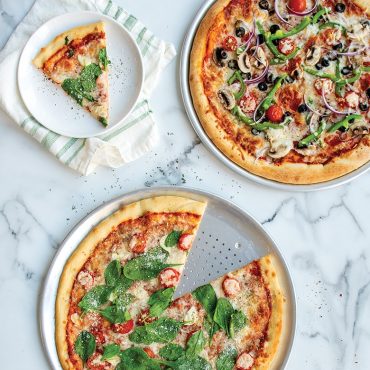 Two Nordic Ware aluminum pizza pans with baked pizzas - one traditional smooth, one perforated crisper pan