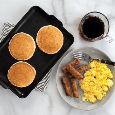 Three golden pancakes on Nordic Ware black non-stick mini griddle with breakfast plate and coffee cup nearby
