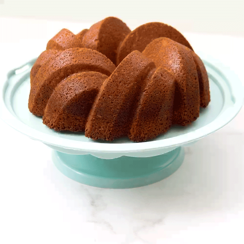 Hand placing clear plastic cover over golden bundt cake on Nordic Ware aluminum cake stand with pedestal base