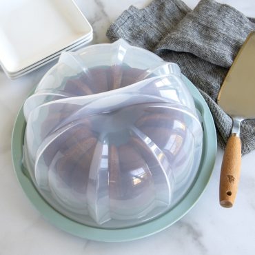 Clear plastic bundt cake keeper with dome lid covering a golden brown bundt cake with fluted design