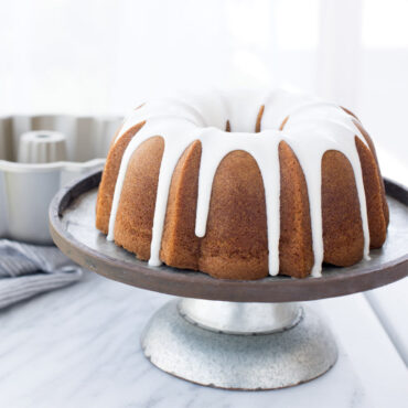 Golden glazed bundt cake on white plate with Nordic Ware silver aluminum bundt pan in background