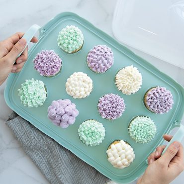 Hands carrying Nordic Ware plastic cupcake carrier with decorated frosted cupcakes, removable lid beside