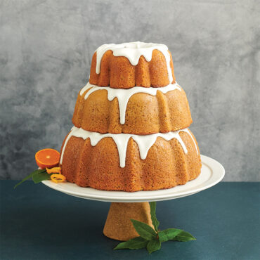 Three golden bundt cakes with white glaze stacked on white pedestal, showcasing Nordic Ware tiered bundt pan set