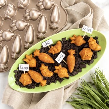 Orange carrot-shaped cakelets with green frosting tops arranged on white platter with chocolate crumb soil, Nordic Ware Garden Patch pan visible in background