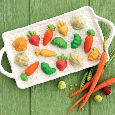 Six glazed vegetable-shaped cakelets from Nordic Ware Garden Patch pan displayed with fresh carrots, pea pods, and radishes