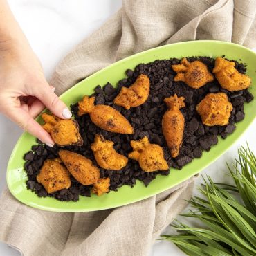 Hand selecting carrot-shaped cakelet from platter of colorful vegetable cakelets with chocolate crumb soil