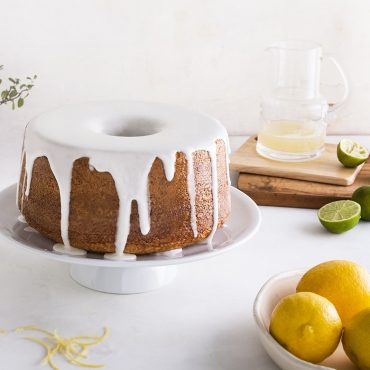Golden brown bundt cake with white glaze on cake stand, fresh citrus and glaze bowl nearby