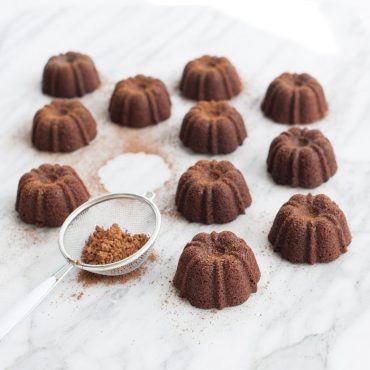 Twelve chocolate mini bundt brownies with fluted ridges arranged on white marble, cocoa powder dusted around